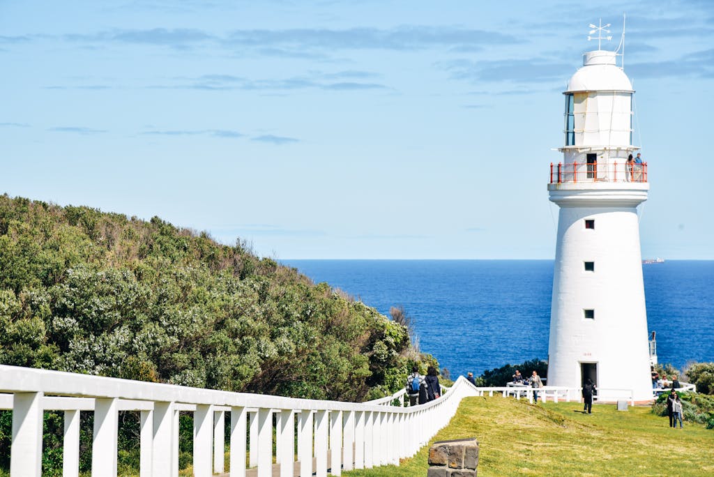 Beautiful lighthouse by the ocean with lush greenery and a clear blue sky.